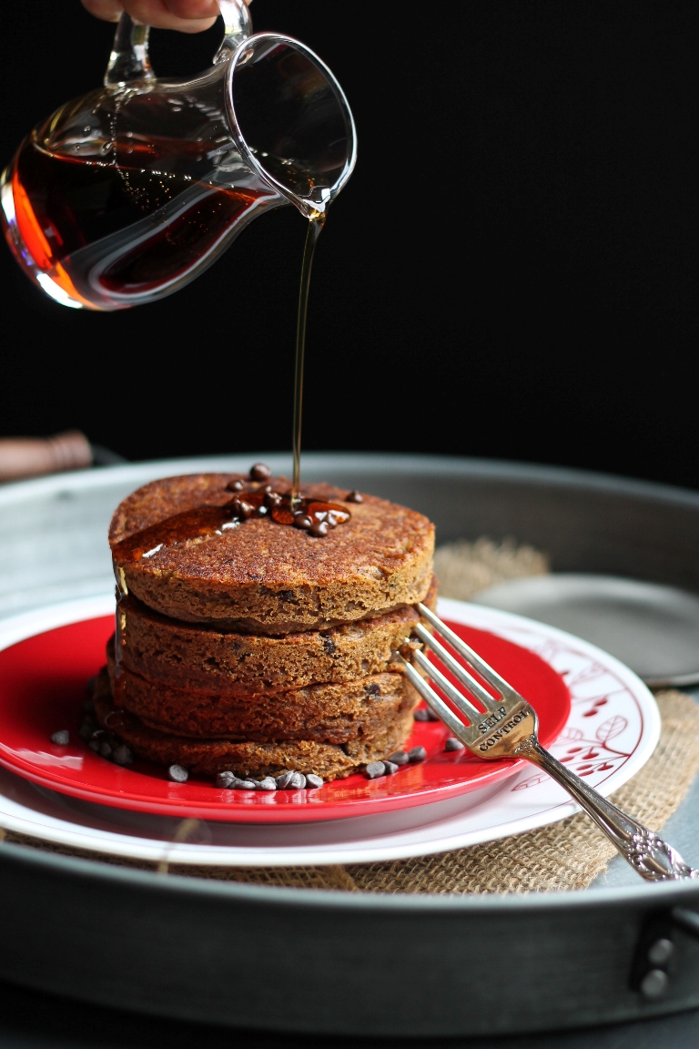 closeup of hand pouring syrup over stack of pancakes with chocolate chips on top
