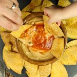 2 hands dipping chips into wood bowl of cheese