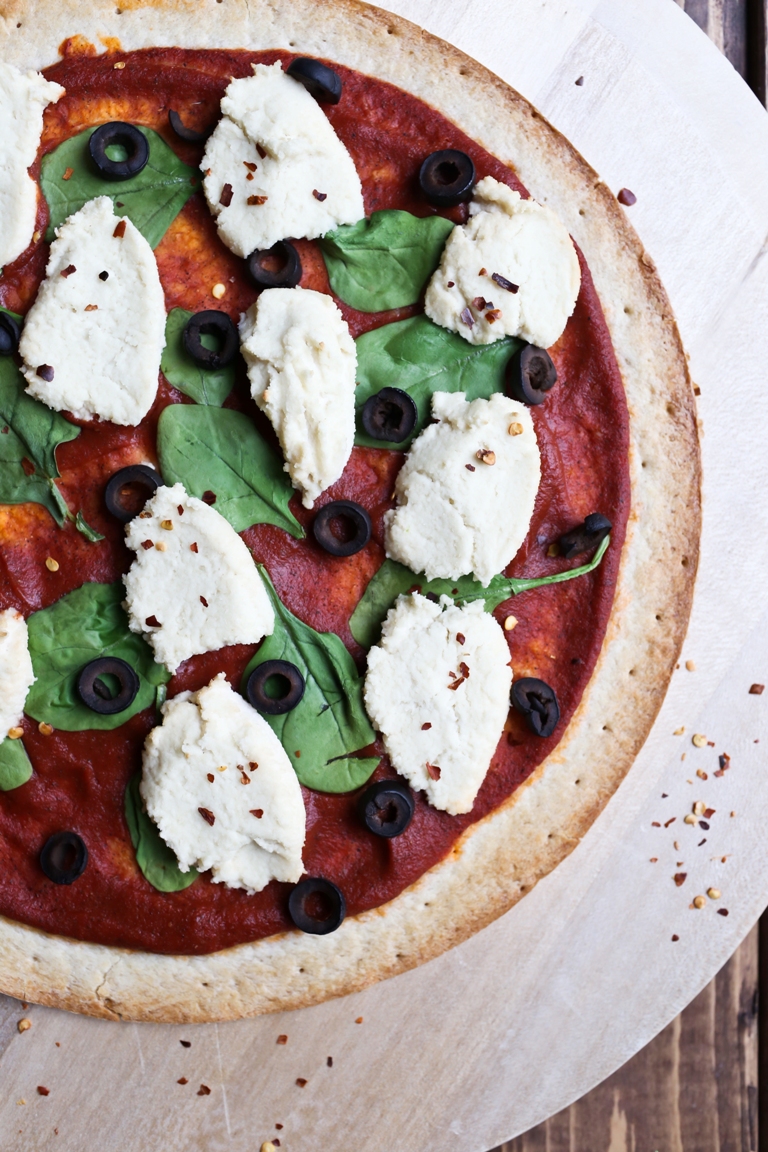 Closeup image of pizza with vegan almond ricotta cheese, spinach and black olives.