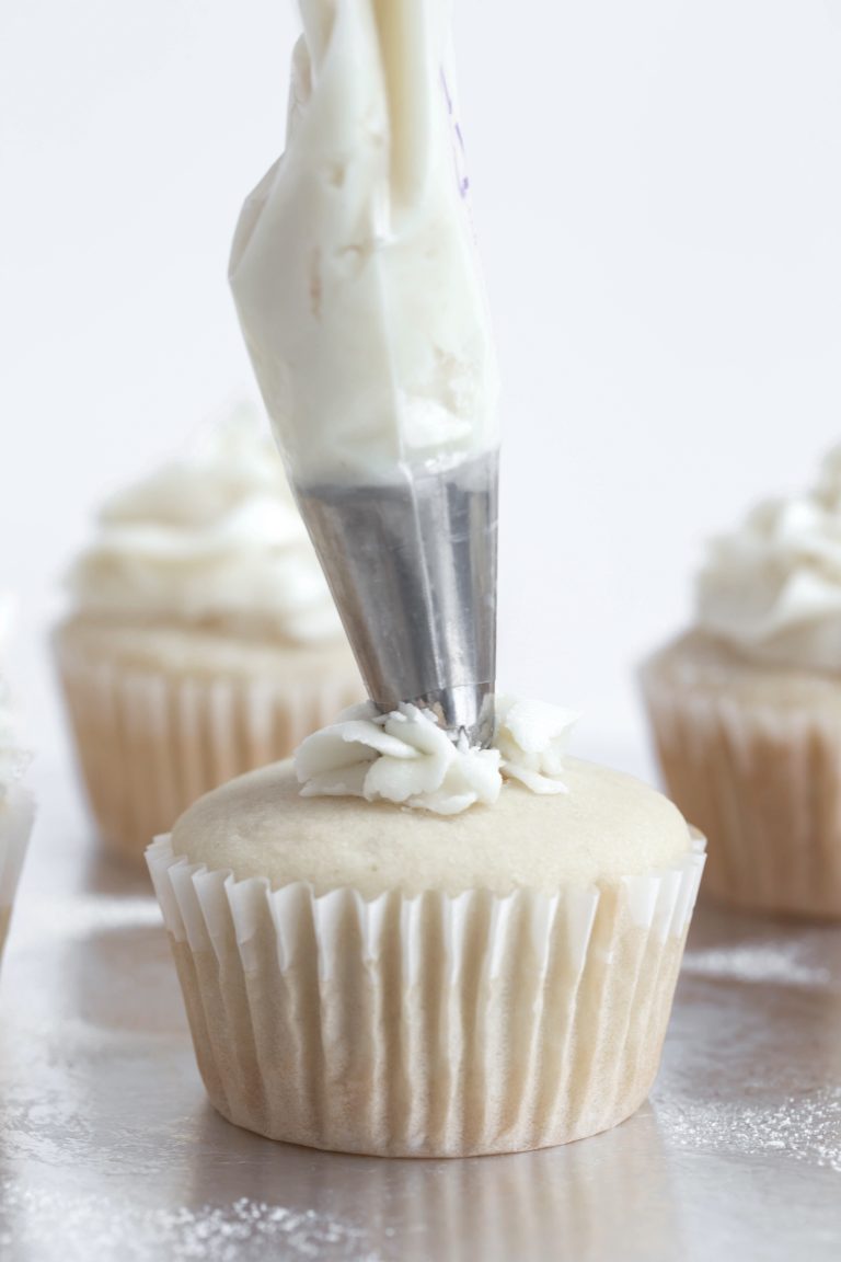 Piping bag with white frosting coming out of the tip onto a white wedding cupcake.