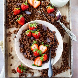bowl of healthy chocolate granola with spoon and fresh strawberries on tray of granola